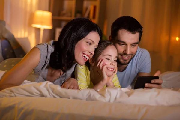 Familia feliz con teléfono inteligente en la cama por la noche —  Fotos de Stock