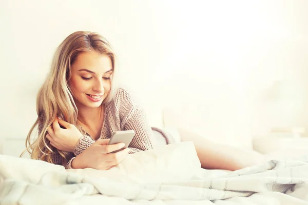 Happy young woman with smartphone in bed at home — Stock Photo, Image