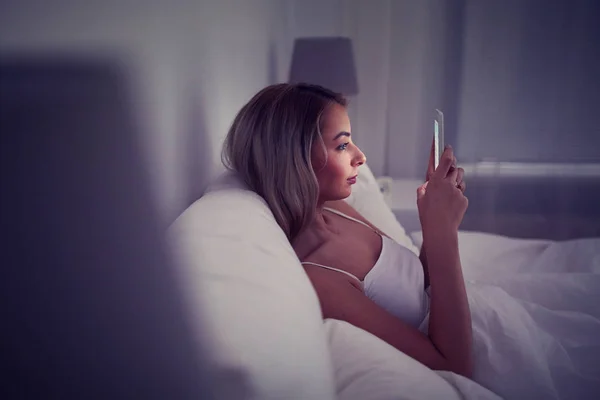 Jonge vrouw met smartphone in bed huis slaapkamer Stockafbeelding