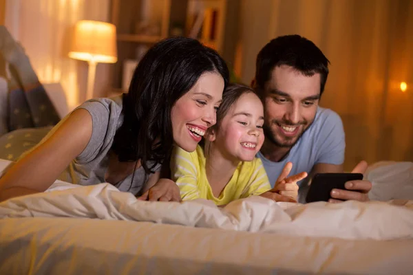 Familia feliz con teléfono inteligente en la cama por la noche —  Fotos de Stock