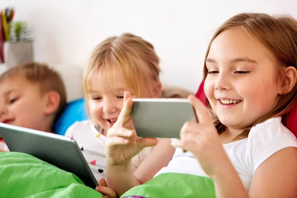 Crianças felizes com tablet pc e smartphone na cama — Fotografia de Stock