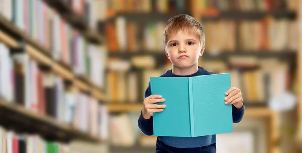 Niezadowolony mały chłopiec czyta książkę nad biblioteką — Zdjęcie stockowe