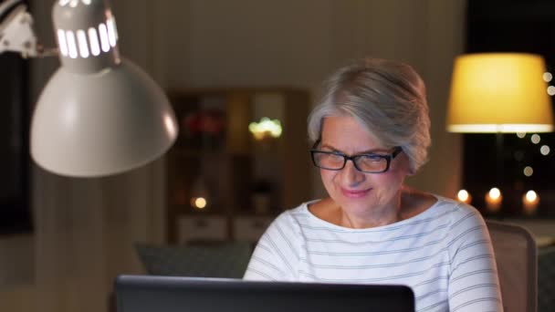 Ευτυχισμένη ηλικιωμένη γυναίκα με φορητό υπολογιστή στο σπίτι το βράδυ — Αρχείο Βίντεο