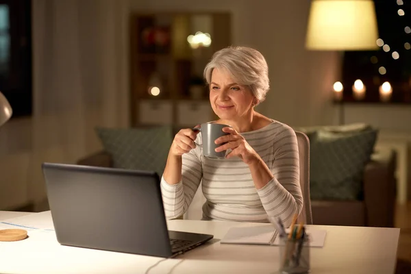 Пожилая женщина с ноутбуком пьет кофе дома — стоковое фото