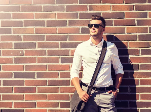 Jeune homme en lunettes de soleil avec sac sur le mur de briques — Photo