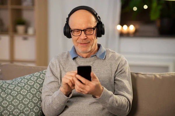 Homem idoso com smartphone e fones de ouvido em casa — Fotografia de Stock