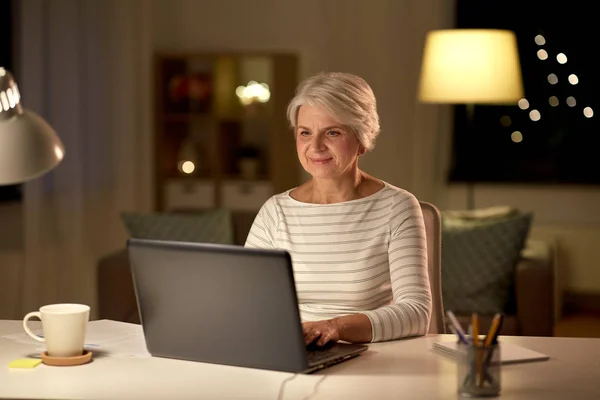 Счастливая пожилая женщина с ноутбуком дома вечером — стоковое фото