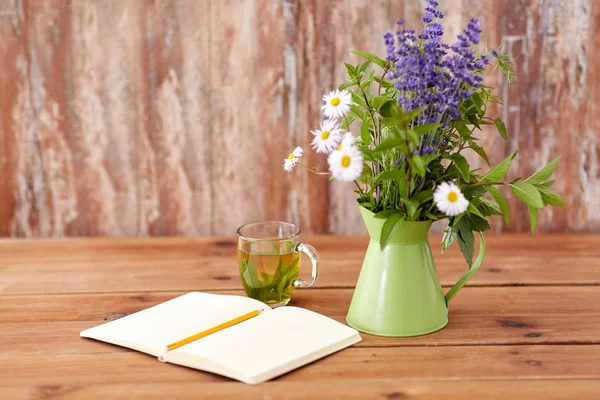 Kräutertee, Notizbuch und Blumen im Krug auf dem Tisch — Stockfoto