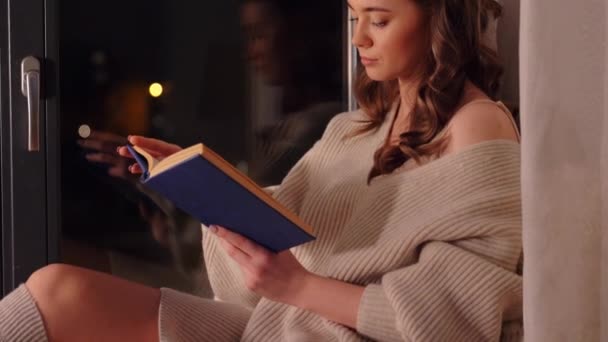 Женщина читает книгу сидя дома на подоконнике — стоковое видео