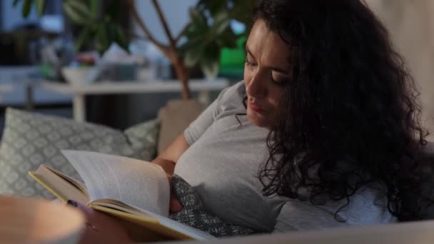 Молодая женщина читает книгу на диване дома — стоковое видео