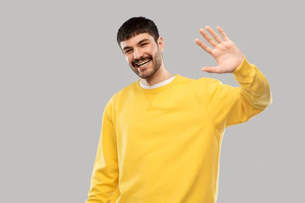 Smiling young man in yellow sweatshirt waving hand — Stock Photo, Image