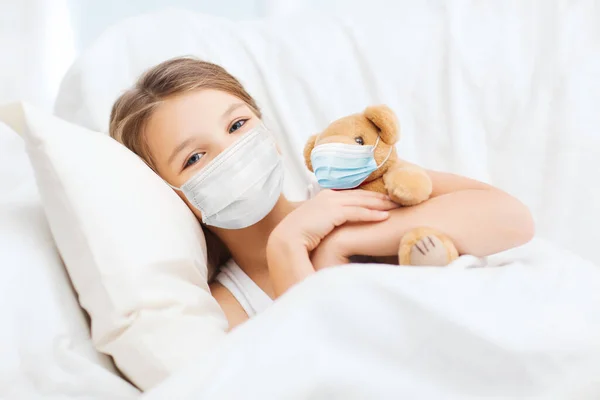 Mädchen mit medizinischer Maske mit Teddybär im Bett — Stockfoto