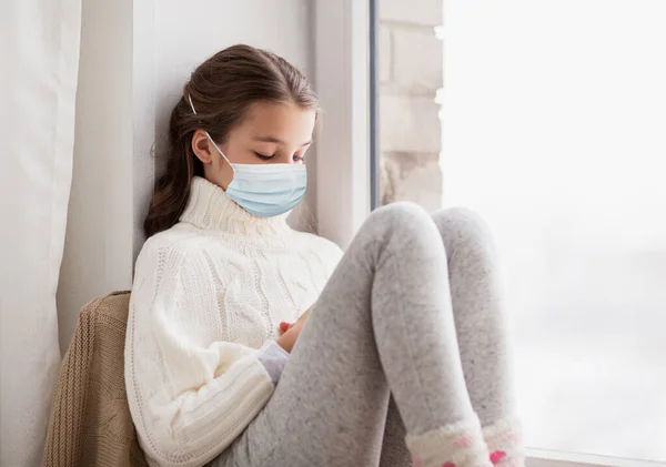 Triste ragazza in maschera medica seduta sul davanzale di casa — Foto Stock