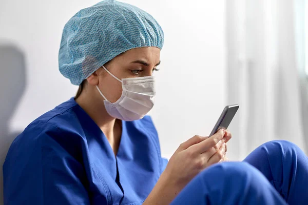 Smutný lékař nebo zdravotní sestra v masce obličeje s smartphonem — Stock fotografie