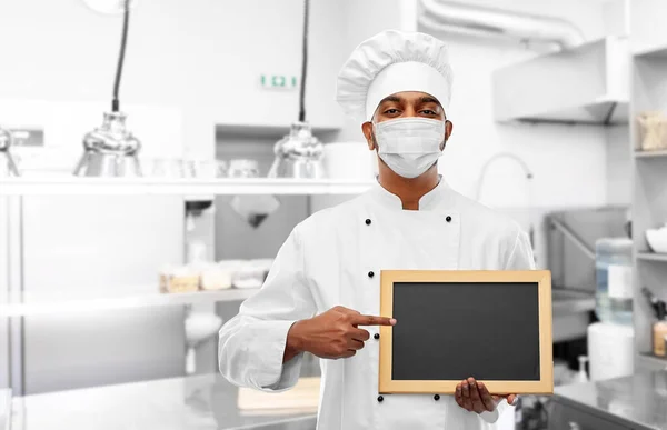 Chef in face mask with chalkboard at kitchen — Stock Photo, Image