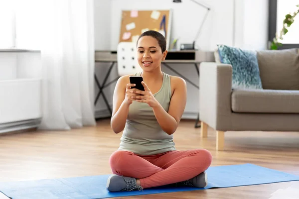 Donna con smartphone si siede sul tappetino da ginnastica a casa — Foto Stock