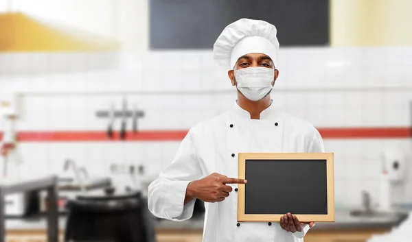 Šéfkuchař v masce s tabulí v kuchyni — Stock fotografie