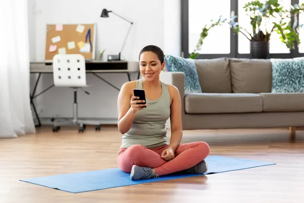 Mujer con teléfono inteligente se sienta en la alfombra de ejercicio en casa — Foto de Stock