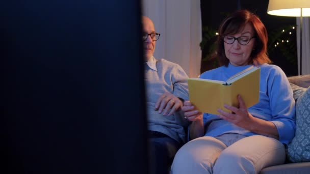 Pareja de ancianos viendo la televisión y leyendo libro en casa — Vídeos de Stock