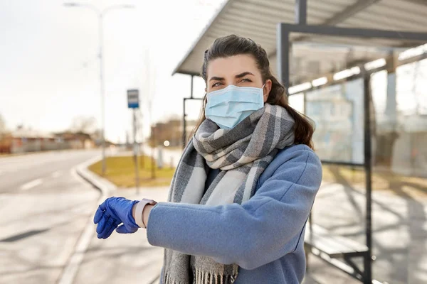 Donna in maschera guardando orologio da polso alla fermata dell'autobus — Foto Stock