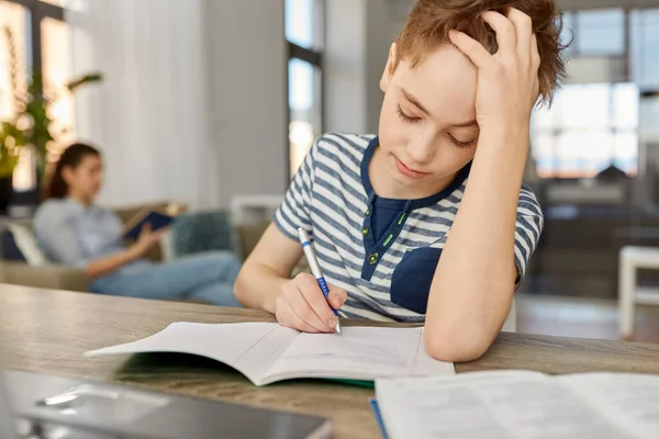 Estudante menino com livro escrevendo para notebook em casa — Fotografia de Stock