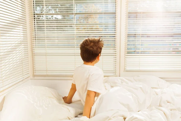Pojken sitter på sängen och tittar ut genom fönster — Stockfoto