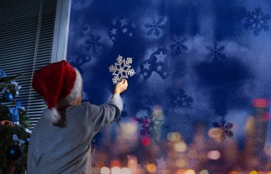 Nice small little boy puts white snowflake on cold frosty winter window glass on Christmas eve evening clipart