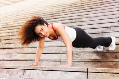 African sportswoman doing push-ups, exercising outdoors on city stairs, looking at camera clipart