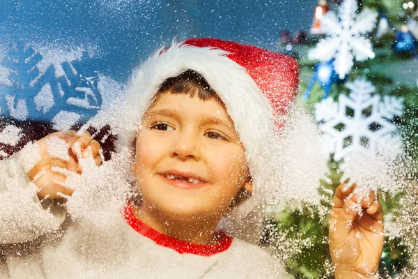 小男孩在圣诞老人的帽子捧着玻璃 看着窗外 在新年前用冰冻的雪花微笑 — 图库照片