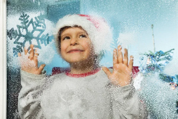 圣诞老人抱着玻璃的小男孩 看着窗外 在圣诞节前带着冰冻的雪花微笑 — 图库照片
