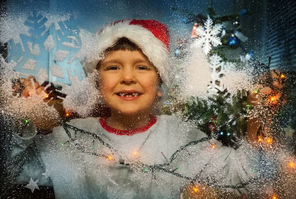 透过冰冷的窗户望着圣诞老人的小男孩在圣诞树和照明附近 — 图库照片