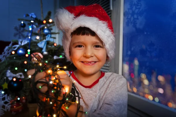 男孩在圣诞老人的帽子笑着微笑 并举行新的一年照明灯即将把它们放在圣诞树上 — 图库照片