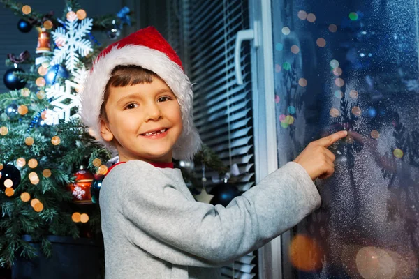 快乐的小男孩指着冬日里冰冷的冬窗 站在圣诞树旁 — 图库照片