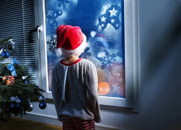 小男孩在圣诞老人的帽子站在寒冷的冬新年窗前 看着夜晚等待圣诞节 — 图库照片