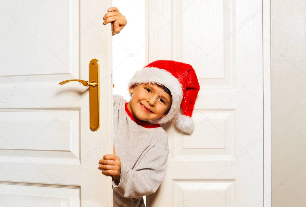 Happy little boy in Santa hat looking curiously behind the white door