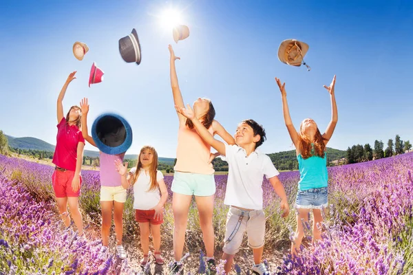 Group of age-diverse kids standing in lavender field and tossing up their hats over blue sky in summer