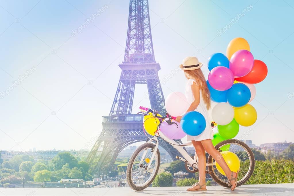 Adorable little girl wearing dress and straw hat, cycling through Paris with colorful balloons bundle