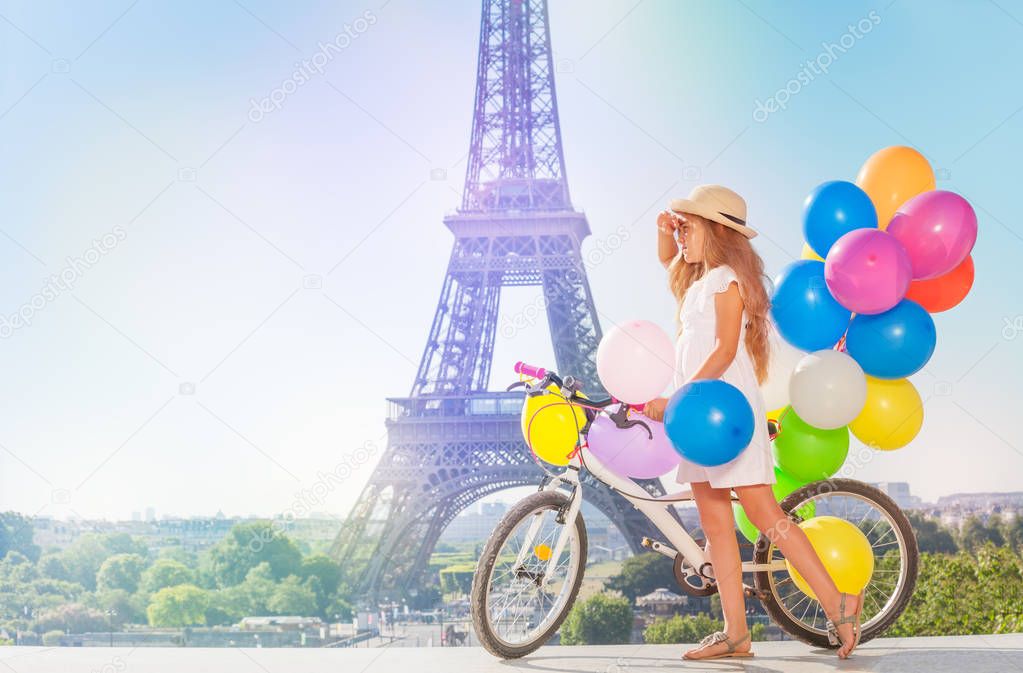 Elegant little girl wearing white dress and straw hat, cycling through Paris with colorful balloons bundle