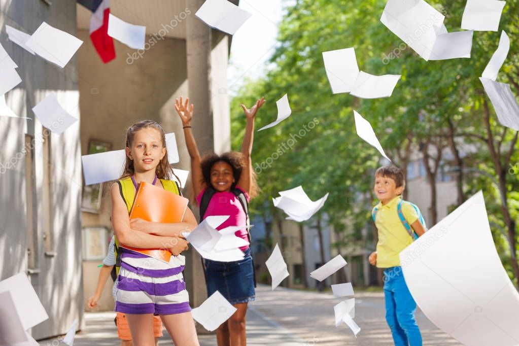 Caucasian girl among group of happy kids throwing papers in the air near the public school with gentle smile