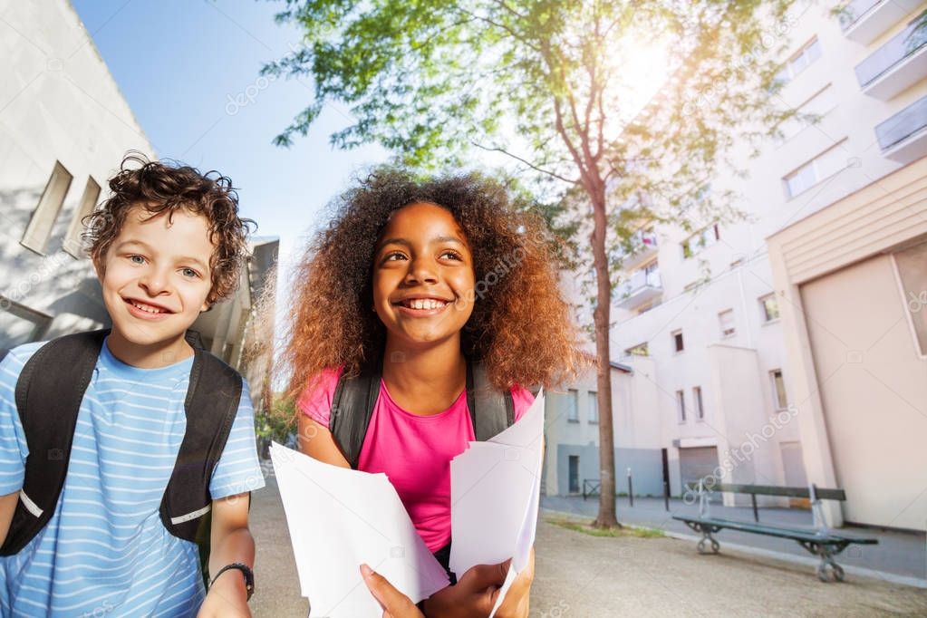 Two school children, Caucasian boy and African girl holding papers looking at camera