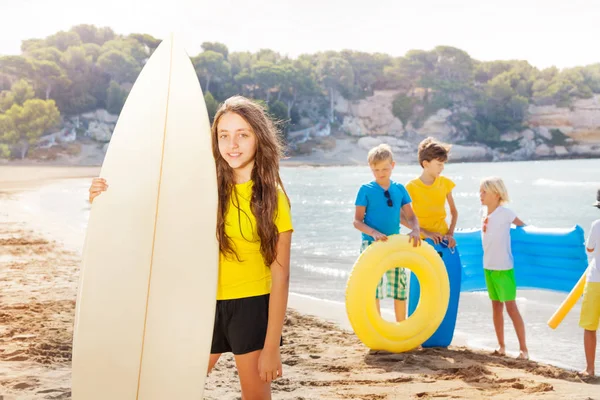 小女孩在海滩站立与冲浪板和她的朋友在后面拿着充气玩具或玻璃活塞 — 图库照片