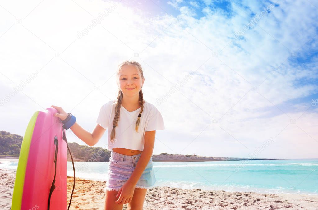 Happy braided girl with body board standing on sandy sunny beach and bending over camera