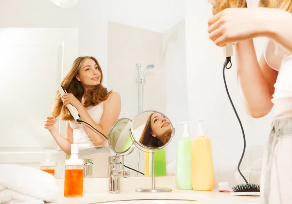 長いウェーブのかかった髪の平らな鉄を使用して バスルームの鏡で見ていると美しい女性 — ストック写真