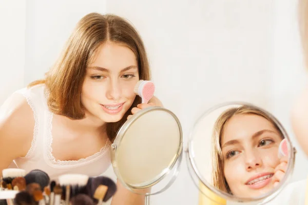 化粧鏡に見られる 皮膚洗浄ブラシを使用して彼女の頬をマッサージ魅力的な若い女性のクローズ アップの肖像画 — ストック写真