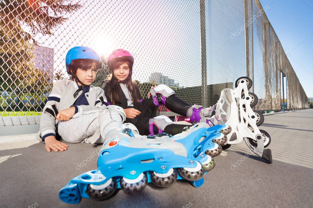 Preteen boy and girl, inline-skaters in safety helmets, sitting on the floor outdoors at stadium