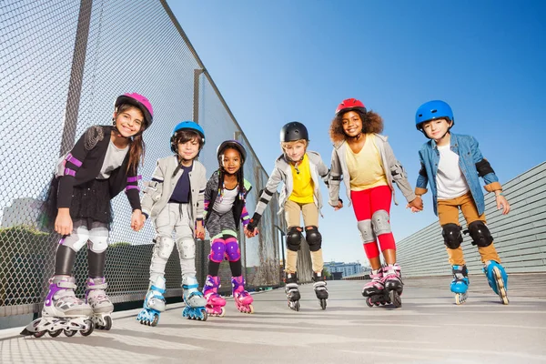 Big group of preteen children, happy in-line skaters in protective gear, standing in a row holding hands at stadium outdoors