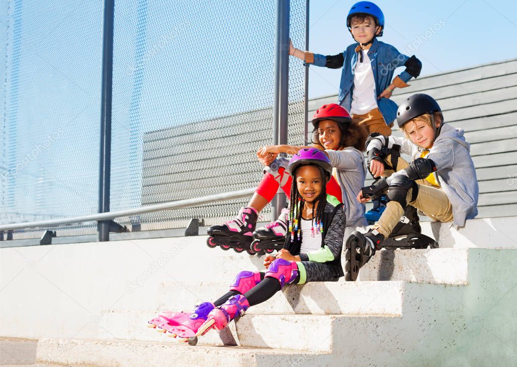 Group of happy preteen kids in roller skates and protective gear, sitting on the steps of stadium outdoors at sunny day