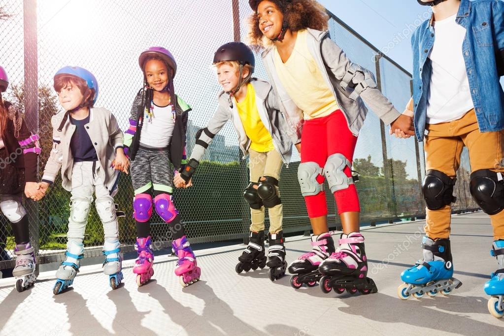 Group of multiethnic children, happy in-line skaters in protective gear, standing in a row holding hands outdoors at sunny day