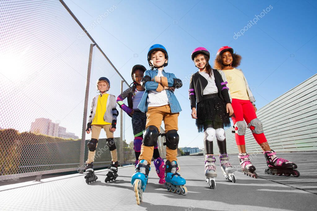 Low-angle view of happy multiethnic children in rollerblades and protective gear posing outdoors at stadium at sunny day
