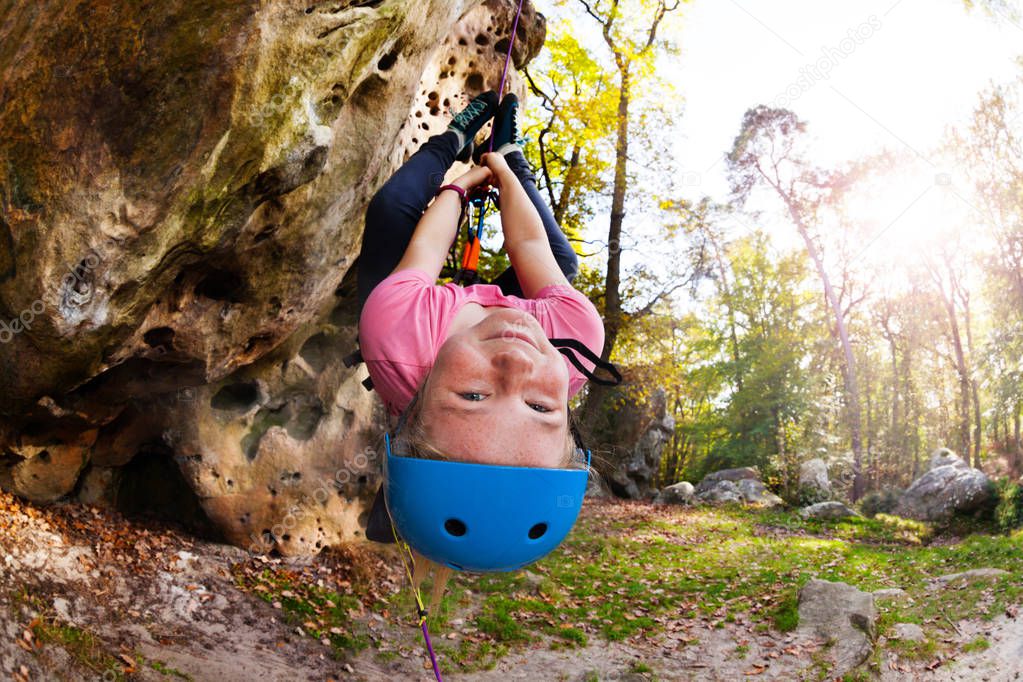 Cute girl in helmet hanging upside-down during rock climbing train outdoors in forest area